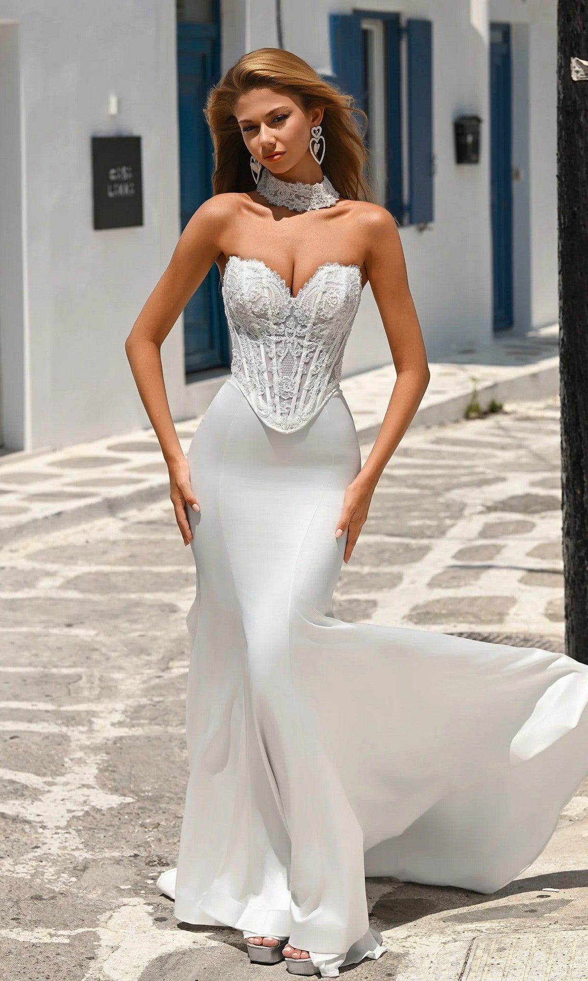 A woman in the Portia and Scarlett PS26086 Long Formal Dress in ivory, featuring a strapless lace corset bodice and satin mermaid skirt, stands on a sunlit stone path with white buildings and blue doors behind her.